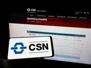 celular com logotipo da CSN na tela e, ao fundo, monitor com gráfico de cotação da ação da CSN